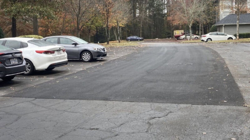 Parking lot surrounded by trees and part of a residential building can be seen in the background. There are multiple parked cars. The parking lot has a large area of asphalt repair where cracks and potholes have been filled.