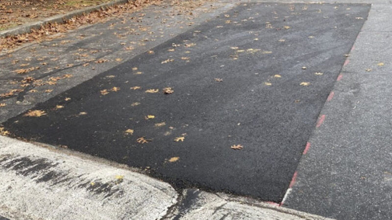 Photo of a road with speed bumps. There is a large area of asphalt repair where there was cracks and potholes filled right in front of the speed bumps.