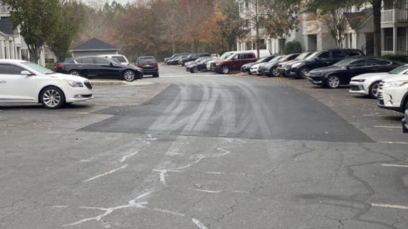 Parking lot with multiple cars parked outside of residential buildings. The parking lot has a large area that has been patched with asphalt to repair cracks and potholes.