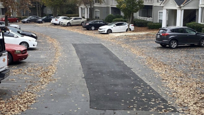 Photo of a parking lot for a residential area with multiple cars parked. There is a large area of asphalt repair in the middle.