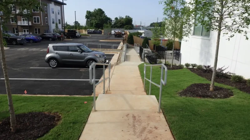 Photo of a handicap ramp that was updated with handrails for ADA compliance. A parking lot and apartment building is off to the left, and there are two tree in grass to the right of the sidewalk.