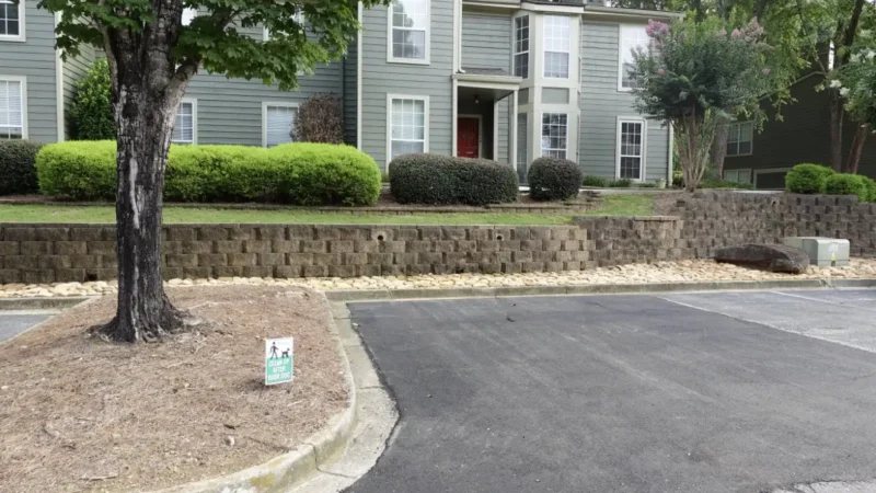 Photo of a parking lot with a residential building in the background. The parking lot has a large area of asphalt patching due to cracks and potholes.