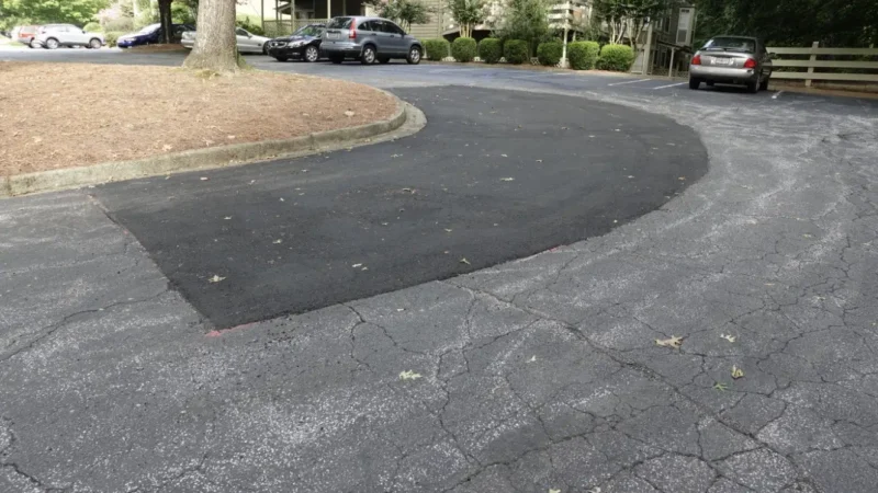 Photo of a parking lot with a building in the background. The entrance to the parking lot has a large area that has been patched with asphalt due to cracks and potholes.