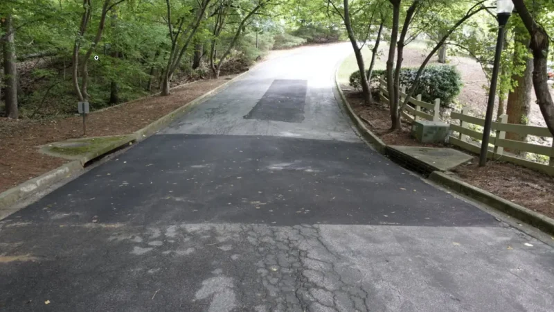 Photo of a road surrounded by trees that you can see two large areas of asphalt patching due to cracks and potholes.