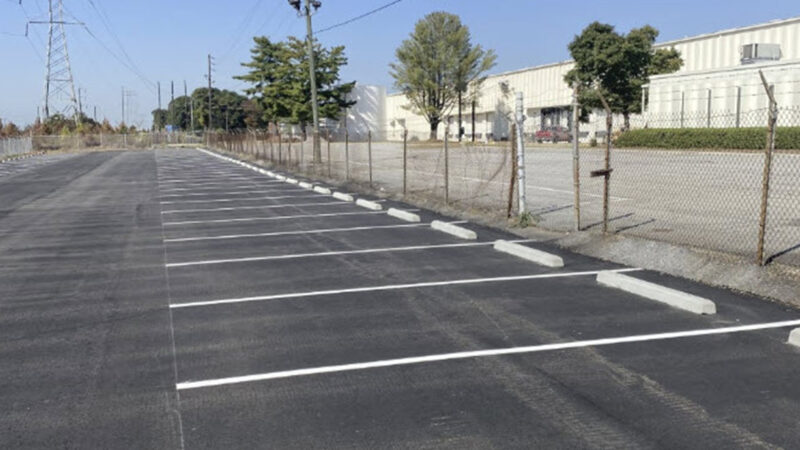 Photo of a parking lot with a fence and warehouse in the background. The parking lot has been sealed and restriped, and there are fresh wheelstops at the front of each parking spot.